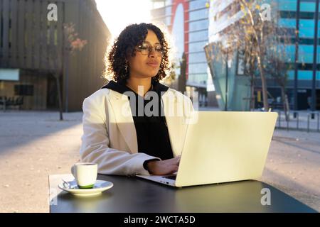A young entrepreneur works on a laptop while seated at an outdoor table at a cafe, with a cup of coffee next to her Stock Photo
