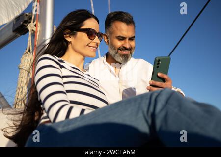 Low angle of smiling couple sitting on yacht and browsing mobile phone while enjoying sunny day against blue sky Stock Photo