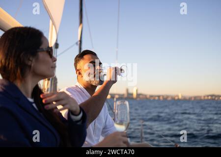 Side couple sitting on yacht in waving ocean and drinking wine while enjoying time together against cloudless sky Stock Photo