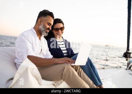 Side view of smiling couple sitting on yacht and browsing laptop while spending time in sea during trip Stock Photo