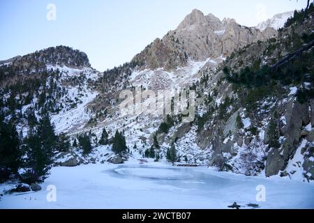 A serene frozen lake set against a backdrop of rugged snowy mountains, with trees emerging from the snow-covered slopes Stock Photo