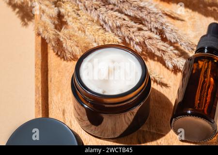 From above close-up of a cream jar and amber dropper bottle on wooden tray with pampas grass, warm lighting Stock Photo