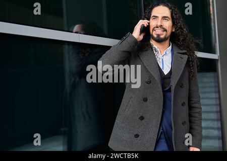 Confident happy mature Latin businessman with long hair looking away while speaking on the mobile phone, outdoors near a modern building Stock Photo