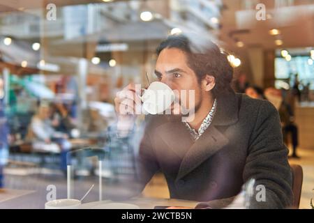 Through window view of thoughtful mature Latin businessman with long hair looking away while drinking coffee in a downtown cafe Stock Photo