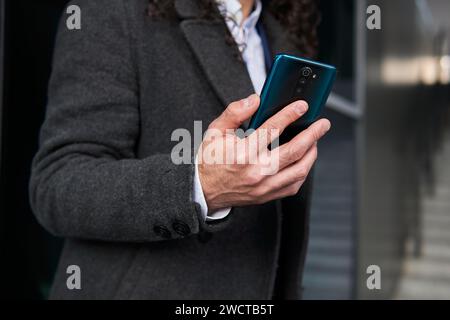 Cropped unrecognizable mature Latin man's hands holding a smartphone, partially visible in a business coat standing on the street Stock Photo
