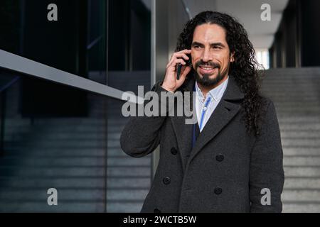 Confident happy mature Latin businessman with long hair looking away while speaking on the mobile phone, outdoors near a modern building Stock Photo