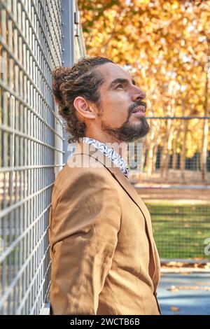 Pensive mature Latin businessman with long hair looking away, leaning against a metal fence in an autumnal urban street Stock Photo