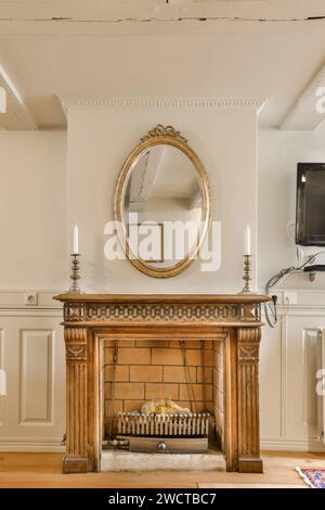 A classic wooden fireplace adorned with an ornate gold-framed oval mirror and flanked by lit candles, set against a neutral wall. Stock Photo
