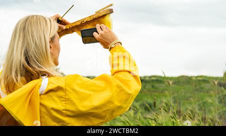 Back view of anonymous female in yellow protective suit looking at screen of mobile phone while standing in field with bee comb on wooden frame and ta Stock Photo