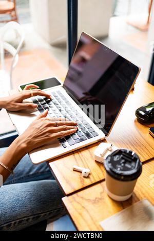 Cropped unrecognizable woman's hands typing on a laptop in a cafe, with a smartphone, earphones, and a coffee cup on the table during remote work in C Stock Photo