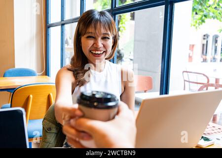 Asian woman smiling and holding a coffee cup in a cafe with laptop during break from remote work in Chiang Mai, Thailand Stock Photo