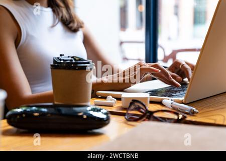Cropped unrecognizable woman's hands typing on a laptop, with a coffee cup and smartphone on the table focused during remote work setting in Chiang Ma Stock Photo