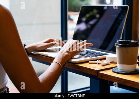 Cropped unrecognizable woman's hands typing on a laptop, with a coffee cup and smartphone on the table focused during remote work setting in Chiang Ma Stock Photo