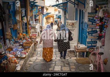 Two Muslim Tunisian women wearing headscarves walk hand-in-hand past a souvenir shop in Rue El Aghalba in the ancient medina, Sousse, Tunisia. The med Stock Photo