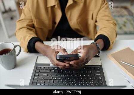 Hands of unrecognizable black man typing on smartphone while sitting in front of laptop at desk Stock Photo
