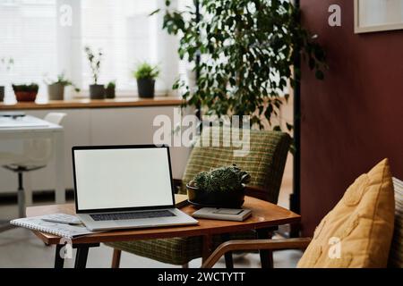 Laptop with white screen mockup placed on table in modern office with greenery and vintage furniture Stock Photo