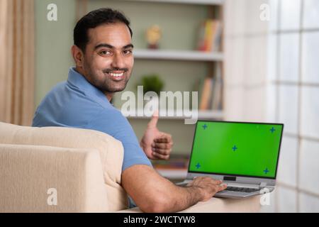 Happy indian man using green screen laptop by showing thumbs up while looking at camera at home - concept of satisfaction, approval and advertisement Stock Photo