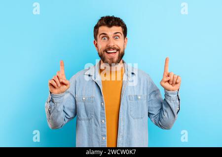 Excited young bearded man pointing fingers up on blue background Stock Photo