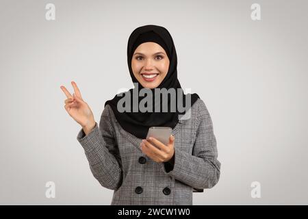 Happy Muslim woman with phone making peace sign Stock Photo