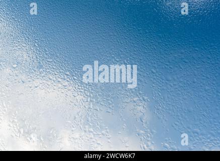 Numerous drops of morning dew on the window glass against the backdrop of a blue sky with clouds. Background Stock Photo