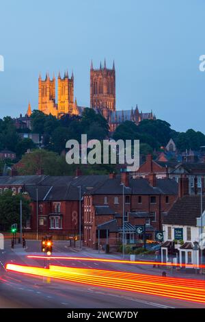 Lincoln, Lincolnshire, England. View along Brayford Way to the illuminated towers of Lincoln Cathedral, dusk, vehicle light trails in foreground. Stock Photo