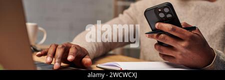 cropped view of african american man with smartphone using laptop while working at home, banner Stock Photo