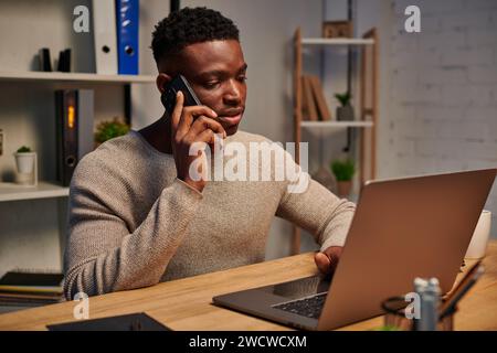 concentrated african american freelancer working from home and talking on smartphone near laptop Stock Photo