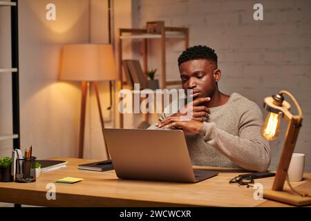 thoughtful creative african american freelancer looking at laptop working from home at night Stock Photo