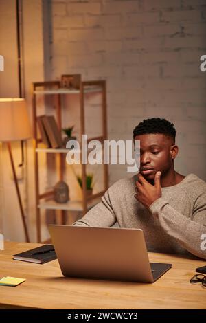 pensive african american freelancer looking at laptop in home office at night, problem solving Stock Photo
