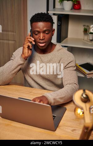 young focused african american freelancer working from home and talking on smartphone near laptop Stock Photo