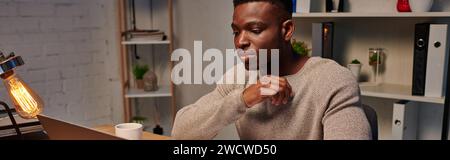 young concentrated african american freelancer working on laptop in evening at home, banner Stock Photo