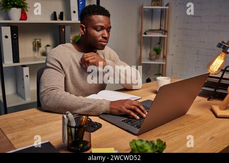 concentrated african american man typing on laptop while working in evening at home, freelancer Stock Photo