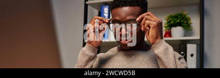 young african american freelancer adjusting eyeglasses while working at home at night, banner Stock Photo