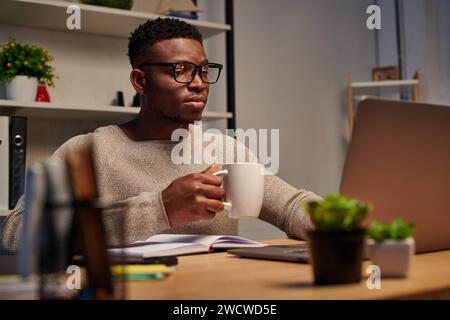 focused african american freelancer in eyeglasses holding coffee cup and working on laptop at home Stock Photo