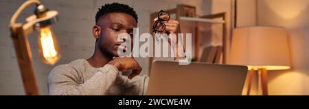 pensive african american freelancer holding eyeglasses looking at laptop in home office, banner Stock Photo