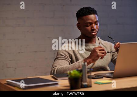 tired african american freelancer holding eyeglasses while sitting at laptop in home office at night Stock Photo