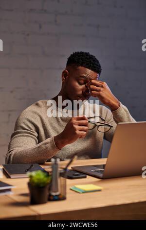 exhausted african american freelancer sitting with eyeglasses near laptop in home office at night Stock Photo
