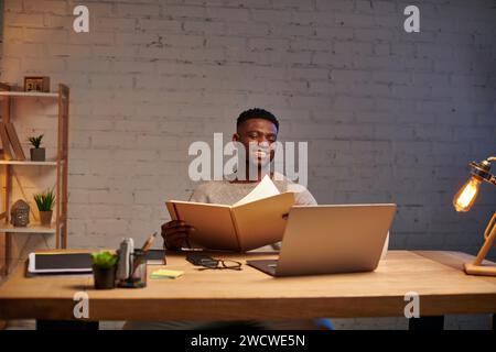 joyful african american freelancer with notebook looking at laptop while working at home at night Stock Photo