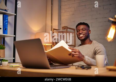 happy african american freelancer with notebook looking at laptop while working at home at night Stock Photo