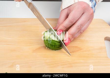 Cutting cucumber into small pieces to make salad. Female hands with a kitchen knife. Homemade food. Stock Photo
