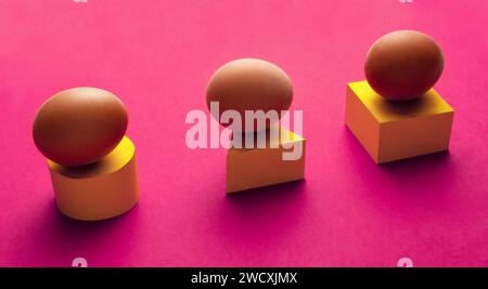 Creative minimal composition of three eggs lying on a yellow cylinder, triangle and cube on a purple background. Stock Photo