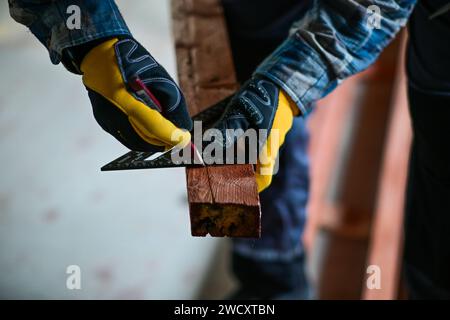 a worker in yellow gloves marks a straight line with a right-angled triangle and a wood pencil to prepare for sawing wood. Stock Photo