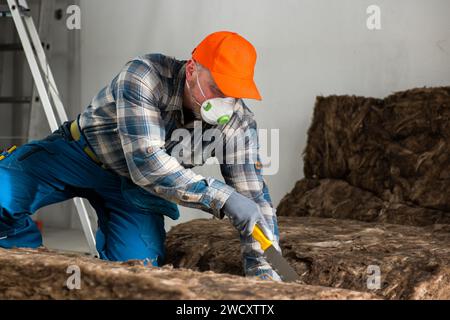 a worker in overalls, gloves and a respirator cuts glass wool with a large knife. Stock Photo