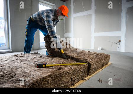 a worker in overalls, gloves and a respirator cuts glass wool with a large knife. Stock Photo