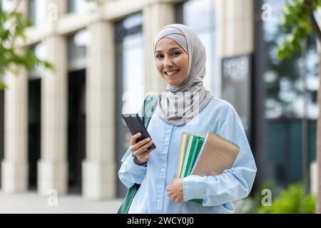 Smiling Arab woman in hijab holding a smartphone and notebook outside an office building, exuding confidence and professionalism. Stock Photo