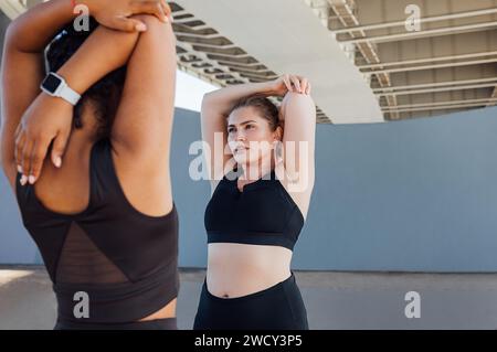 Two plus-size women stretch their hands while standing under a bridge. Young woman warming up hands while standing outdoors with her friend. Stock Photo