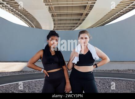 Portrait of two women in black fitness wear with towels on their necks. Young sportswomen are relaxing outdoors. Stock Photo