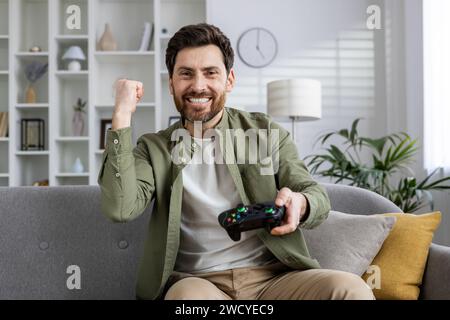 Smiling man sitting on sofa, triumphantly fist-pumping as he enjoys playing a console game in a cozy living room setup. Stock Photo