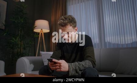 Smiling man sitting on couch at home typing text message by smartphone. Male person relaxing at home. Communicating with family and friends Stock Photo