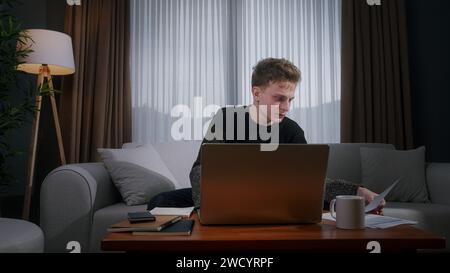Focused blonde man working from home using with laptop while sitting on sofa at home. Focused millennial male looking at laptop, taking note on notebo Stock Photo
